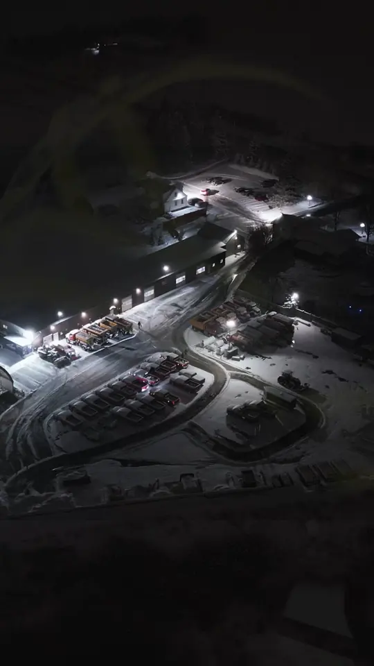 Night aerial of BP Landscaping's snow-covered yard with plow trucks staged under floodlights.